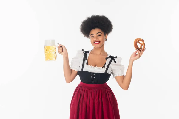 Gioiosa cameriera afroamericana in costume bavarese in posa con boccale di birra e pretzel su bianco — Foto stock