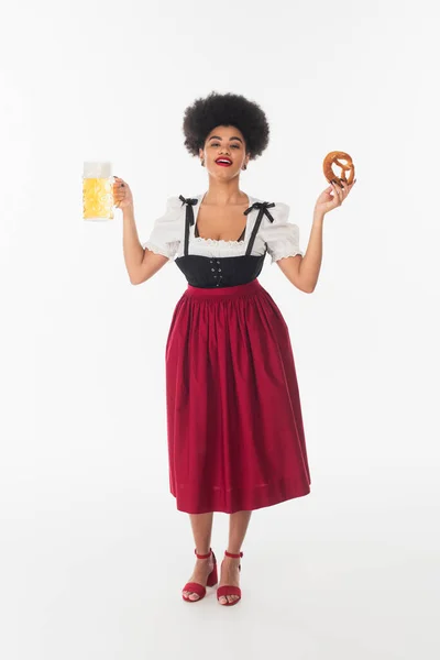 Fröhliche afrikanisch-amerikanische Kellnerin im Oktoberfestkostüm mit Bierkrug und Brezel auf weißem Grund — Stockfoto
