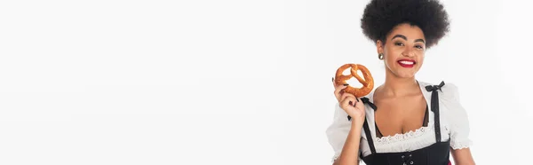 Feliz africano americano oktoberfest camarera en dirndl posando con sabroso pretzel en blanco, bandera - foto de stock