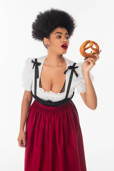 Amazed african american bavarian waitress in oktoberfest attire looking at tasty pretzel on white — Stock Photo