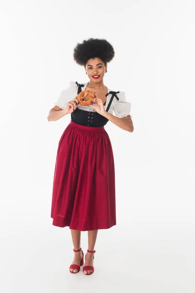 African american bavarian waitress in elegant dirndl with delicious pretzel on white, full length — Stock Photo