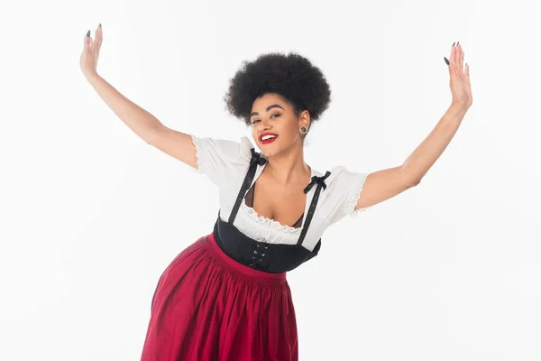 Lively african american waitress in traditional bavarian costume dancing with raised hands on white — Stock Photo