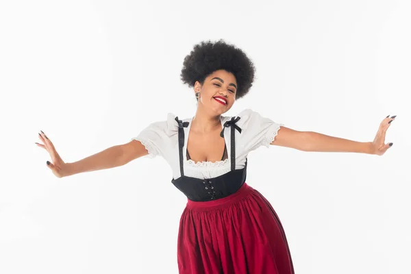 Joyful african american waitress in traditional bavarian costume with outstretched hands on white — Stock Photo