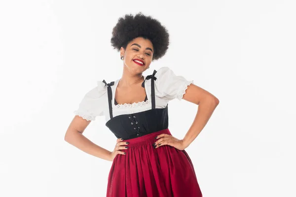Allegra cameriera afroamericana in costume bavarese con le mani sui fianchi su bianco, oktoberfest — Foto stock