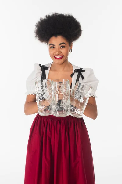 Joyful african american bavarian waitress in dirndl holding empty beer mags and smiling on white — Stock Photo