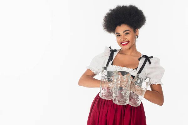 Encantadora camarera bavariana afroamericana en traje oktoberfest celebración de revistas de cerveza vacías en blanco - foto de stock