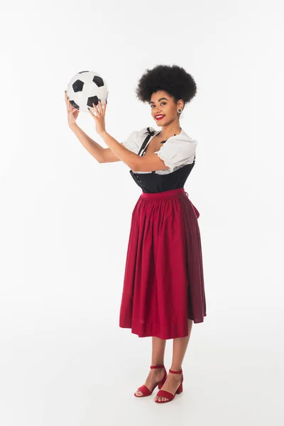 Souriant serveuse bavaroise afro-américaine en robe de dirndl authentique tenant ballon de football sur blanc — Photo de stock