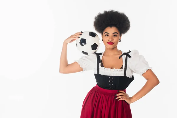Fröhliche afrikanisch-amerikanische Kellnerin im Dirndl posiert mit Fußball und Hand auf Hüfte auf weiß — Stockfoto