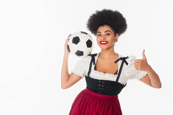 Gioiosa cameriera afroamericana oktoberfest in Dirndl tenendo palla da calcio e mostrando come su bianco — Foto stock