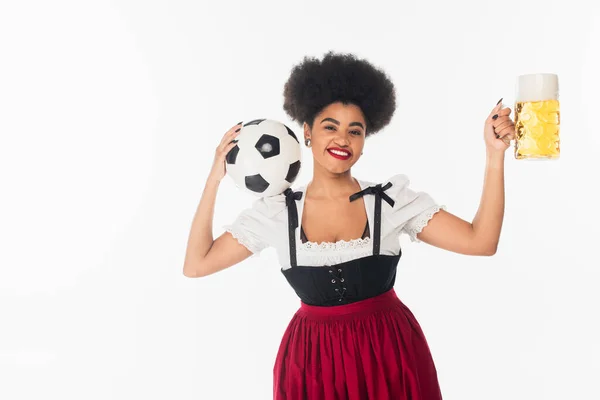 Gioiosa cameriera afroamericana in Dirndl bavarese in posa con pallone da calcio e tazza di birra su bianco — Foto stock
