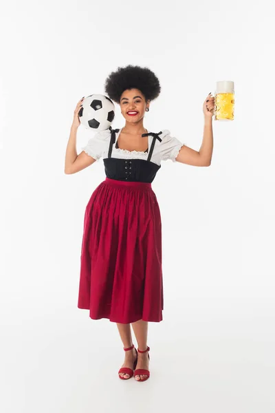Eccitata cameriera afroamericana in costume oktoberfest con boccale di birra e pallone da calcio su bianco — Foto stock