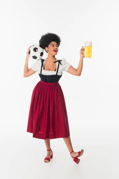 Joyful african american bavarian waitress in authentic attire with soccer ball and beer mug on white — Stock Photo
