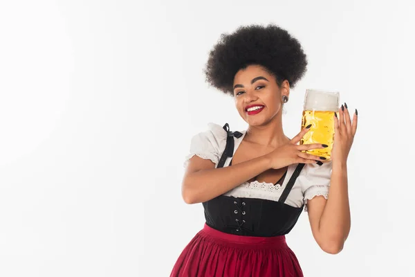 Cheerful african american waitress in oktoberfest outfit holding mug of beer with foam on white — Stock Photo