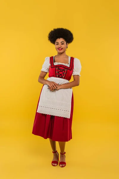 Joyful african american bavarian waitress in oktoberfest costume standing on yellow, full length — Stock Photo