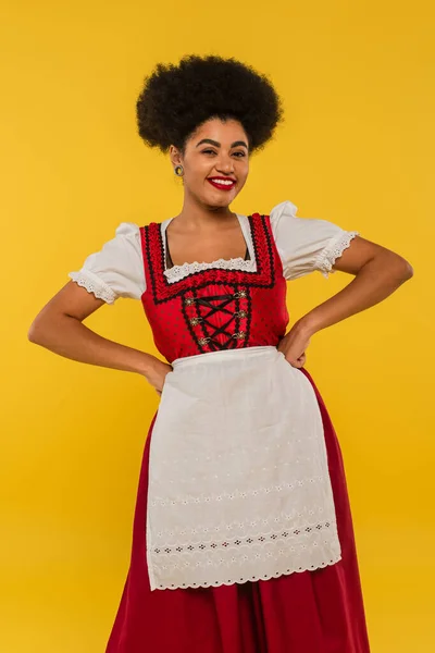Cheerful african american bavarian waitress in dirndl holding hands on waist on yellow, oktoberfest — Stock Photo