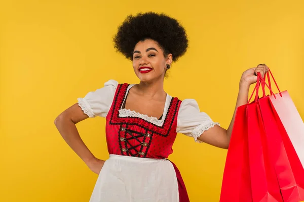 Cheerful african american oktoberfest waitress posing with hand on hip and shopping bags on yellow — Stock Photo