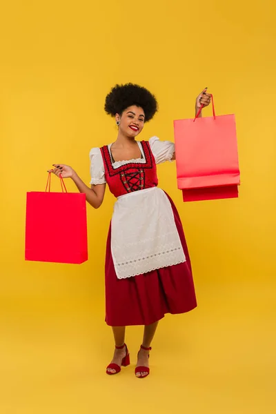 Heureuse serveuse bavaroise afro-américaine en robe traditionnelle debout avec des sacs à provisions sur jaune — Photo de stock