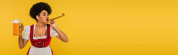 African american bavarian waitress with mug of craft beer blowing in party horn on yellow, banner — Stock Photo