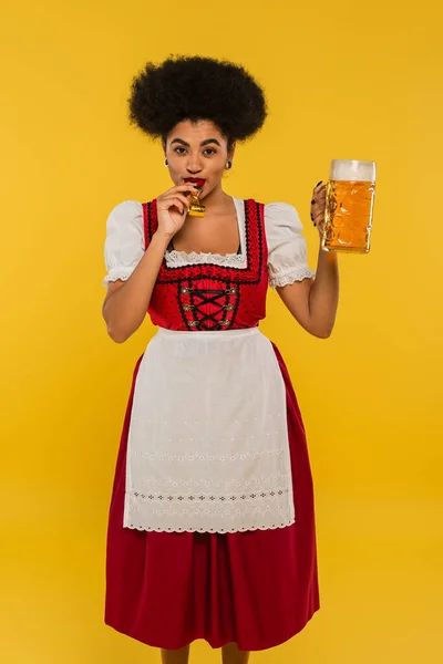 Cheerful african american oktoberfest waitress with mug of beer blowing in party horn on yellow — Stock Photo