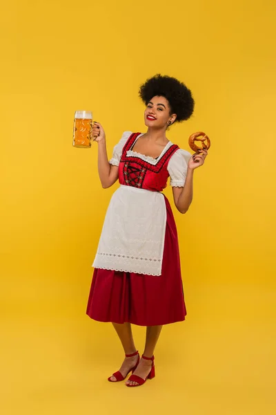 Joyful african american waitress in bavarian costume with beer mug and delicious pretzel on yellow — Stock Photo