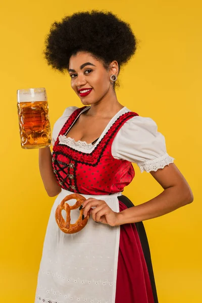 Encantador afroamericano oktoberfest camarera con taza de cerveza y pretzel sonriendo a la cámara en amarillo - foto de stock