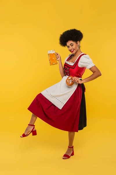 Happy african american waitress in oktoberfest outfit with pretzel and beer on yellow, full length — Stock Photo
