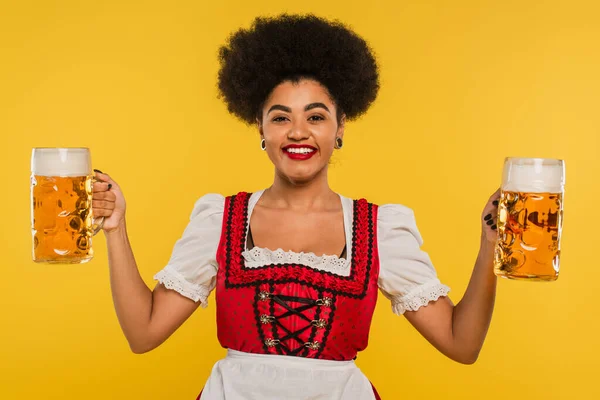 Cheerful african american oktoberfest waitress with mugs of craft beer looking at camera on yellow — Stock Photo