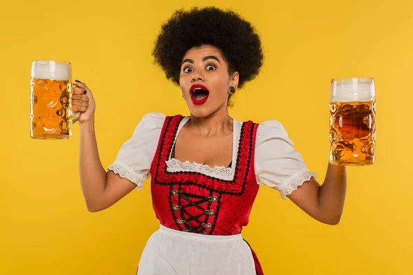 Thrilled african american bavarian waitress with open mouth standing with full beer mags on yellow — Stock Photo