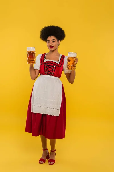 Africano americano camarera en oktoberfest dirndl celebración cerveza tazas y sonriendo a cámara en amarillo - foto de stock