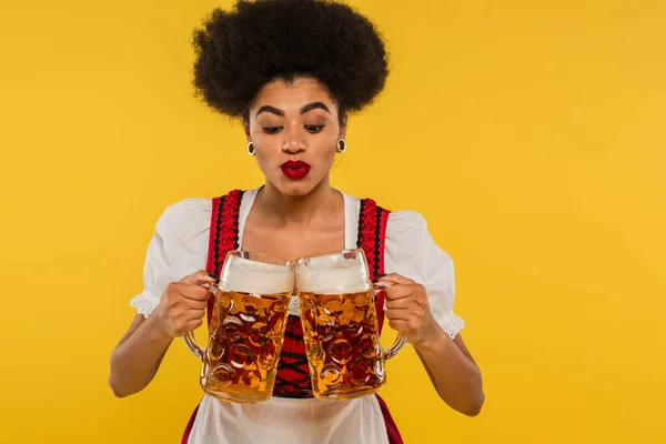 Young african american oktoberfest waitress in bavarian costume clinking mugs with beer on yellow — Stock Photo