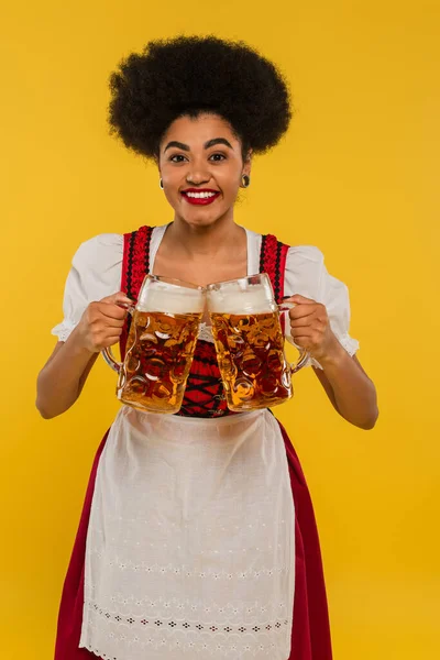 Alegre africana americana garçonete em oktoberfest traje clinking canecas de cerveja e sorrindo no amarelo — Fotografia de Stock