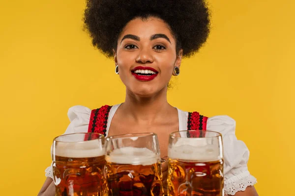 Fröhliche afrikanisch-amerikanische bayerische Kellnerin blickt in die Kamera neben Bechern mit Craft Beer auf gelb — Stockfoto