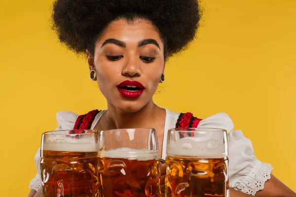 Bastante africano americano oktoberfest camarera en traje bavariano mirando tazas de cerveza completa en amarillo - foto de stock