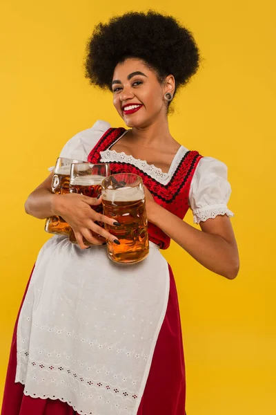 Allegra cameriera afroamericana oktoberfest in abbigliamento autentico che tiene tre tazze di birra sul giallo — Stock Photo