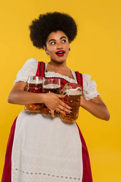Alegre africano americano bavarian camarera en dirndl celebración cerveza tazas y mirando hacia otro lado en amarillo - foto de stock