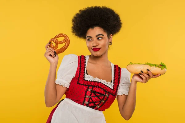 Happy african american oktoberfest waitress in traditional dress with hot dog and pretzel on yellow — Stock Photo