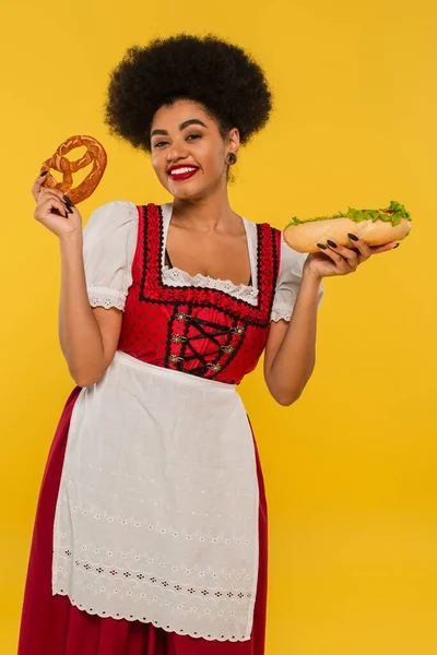 Pretty african american oktoberfest waitress with hotdog and pretzel smiling at camera on yellow — Stock Photo