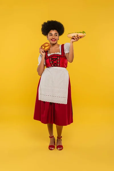 Pretty, happy african american bavarian waitress in dirndl posing with hot dog and pretzel on yellow — Stock Photo