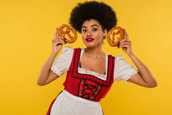 Smiling african american oktoberfest waitress in dirndl showing delicious pretzels on yellow — Stock Photo