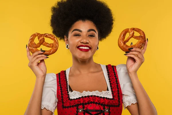 Joyful african american oktoberfest waitress in bavarian dirndl holding tasty pretzels on yellow — Stock Photo