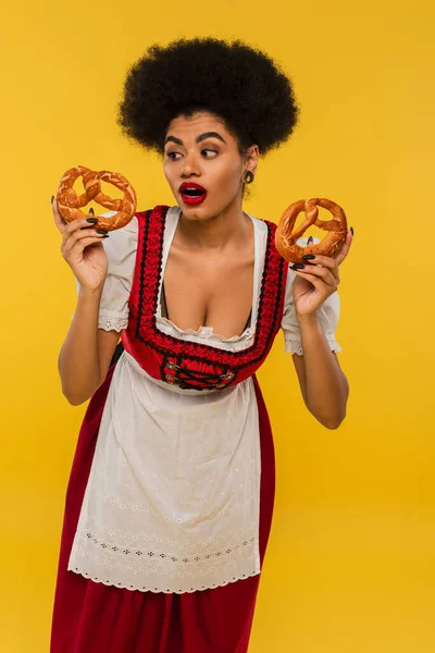 Amazed african american waitress in oktoberfest outfit holding delicious pretzels on yellow — Stock Photo