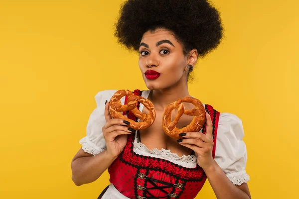 Charmante afrikanisch-amerikanische Oktoberfest-Kellnerin mit leckeren Brezeln blickt in die Kamera auf gelb — Stockfoto