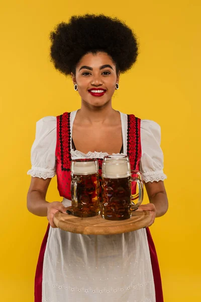 Happy african american waitress in oktoberfest dress serving bavarian beer in mugs on tray on yellow — Stock Photo