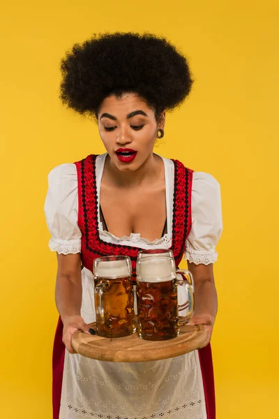African american bavarian waitress in oktoberfest outfit with beer mugs on wooden tray on yellow — Stock Photo