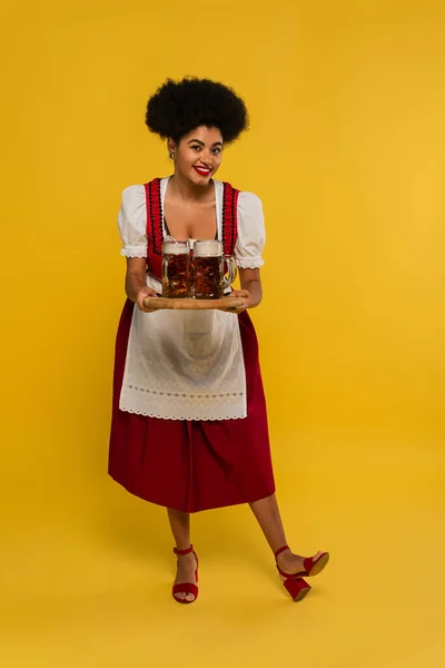 Encantadora oktoberfest afroamericana camarera con tazas de cerveza en bandeja de madera sonriendo en amarillo - foto de stock