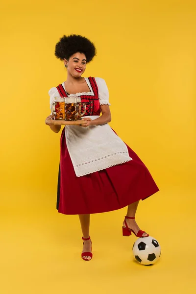 Felice afro americano oktoberfest cameriera con tazze di birra su vassoio di legno vicino al pallone da calcio su giallo — Foto stock