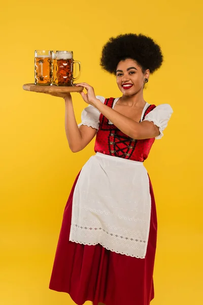Joyful african american bavarian waitress in dirndl serving beer mugs on wooden tray on yellow — Stock Photo