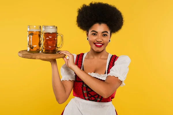 Happy african american bavarian waitress in authentic dress with beer mugs on wooden tray on yellow — Stock Photo