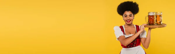 Sorridente cameriera afroamericana oktoberfest con boccali di birra su vassoio di legno su giallo, banner — Foto stock