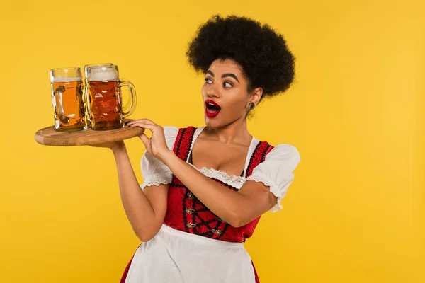Amazed african american oktoberfest waitress holding wooden tray with beer mugs on yellow, banner — Stock Photo